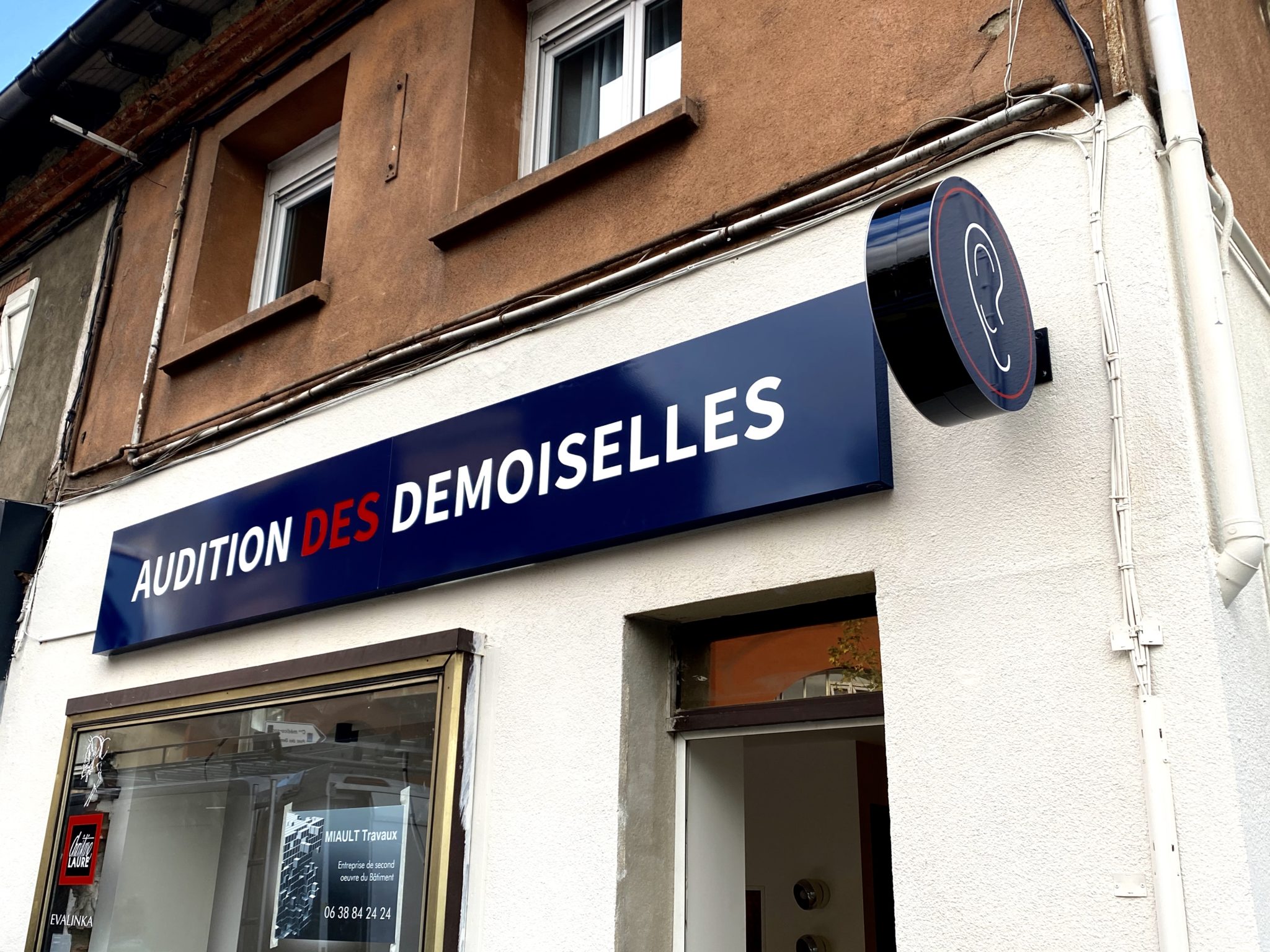 Façade Audition des Demoiselles avec enseigne lumineuse bleue et enseigne drapeau ronde à Toulouse