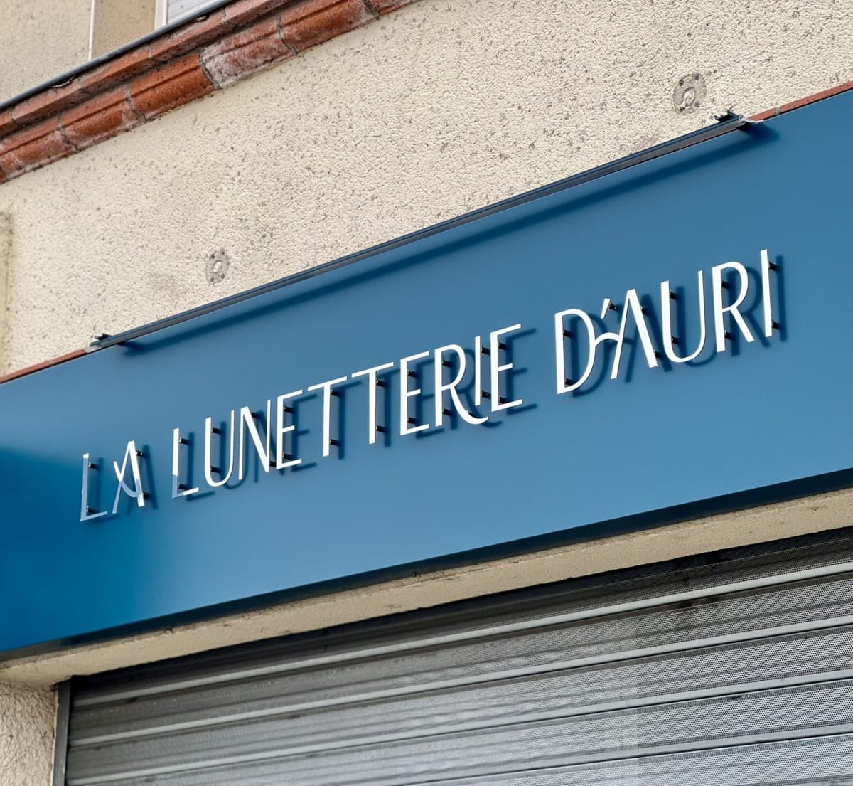 Enseigne de la boutique La Lunetterie d’Auri avec lettres découpées blanches sur bandeau aluminium bleu